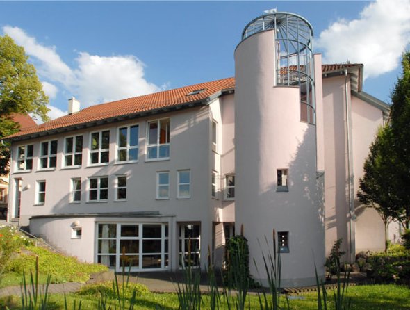 Helles mehrstöckiges Gebäude mit roten Ziegeldächern und einem runden Treppenturm mit Glasaufsatz. Vor dem Haus befinden sich eine Grünfläche, Pflanzen und ein kleiner Weg unter blauem Himmel.