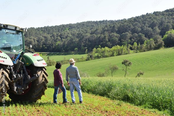 Zwei Personen stehen neben einem Traktor auf einem Feld und blicken über eine grüne Hügellandschaft mit Wald im Hintergrund. Zwei Personen stehen neben einem Traktor auf einem Feld und blicken über eine grüne Hügellandschaft mit Wald im Hintergrund.