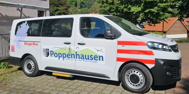 Der Bürgerbus der Gemeinde Poppenhausen (Wasserkuppe) Weißer Bürgerbus der Gemeinde Poppenhausen mit Gemeindelogo und roter Sicherheitsmarkierung, geparkt vor einem Gebäude.