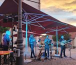 Live-Musik bei einer Open-Air-Veranstaltung: Eine Band mit Gesang, Gitarre und Blasinstrumenten spielt auf einem Dorfplatz unter Sonnenschirmen während der Abenddämmerung.